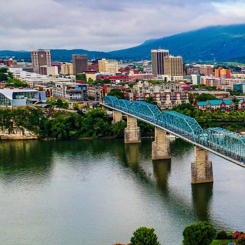 Aerial view of a city skyline with a blue bridge over a river, surrounded by green trees and mountains in the background.