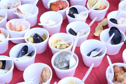 A variety of seafood served in small white cups with plastic spoons on a red tablecloth.