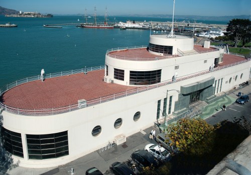 A modern building with a curved roof, overlooking a harbor with boats and a clear blue sky.