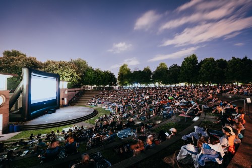 A large outdoor movie screening with a crowd seated on the grass under a night sky, surrounded by trees.