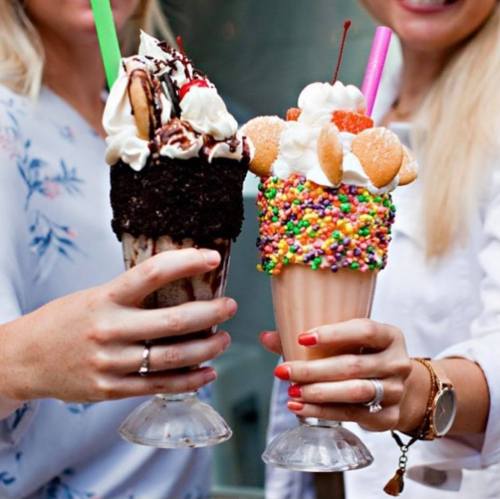 Two hands hold colorful milkshakes topped with whipped cream, cookies, and sprinkles, with bright straws.