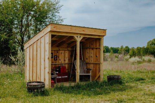 A small wooden shed in a grassy field, surrounded by trees and barrels, under a clear blue sky.