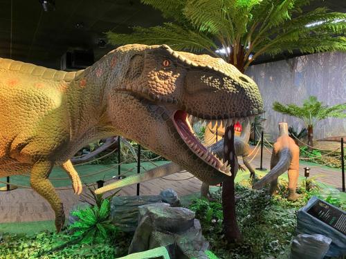 A lifelike dinosaur model in a lush, prehistoric-themed exhibit with greenery and other dinosaur figures.