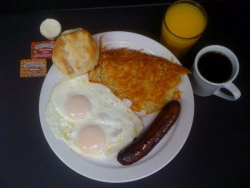 A breakfast plate with two fried eggs, hash browns, sausage, a biscuit, butter, jam, coffee, and orange juice.