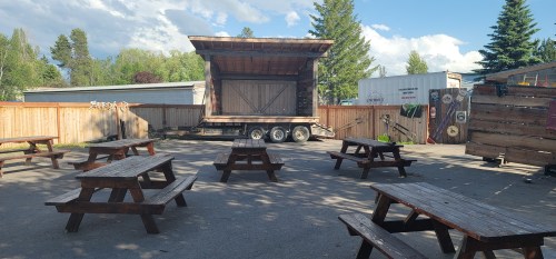 Outdoor venue with wooden picnic tables and a stage on a trailer, surrounded by trees and a wooden fence.