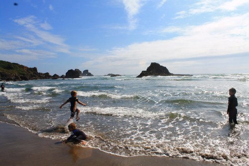 Children playing in the surf at a beach, with rocky formations in the background and a bright blue sky.