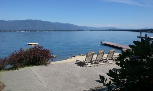 A serene lakeside view with lounge chairs, a dock, and mountains in the background under a clear blue sky.