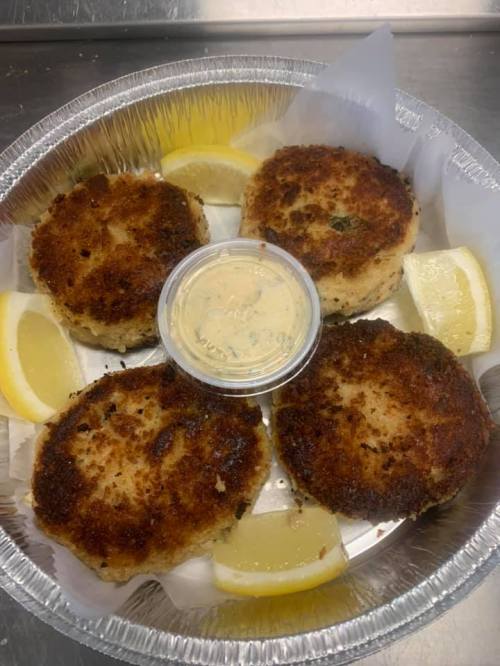 Four golden-brown crab cakes on a platter, served with lemon wedges and a small container of dipping sauce.