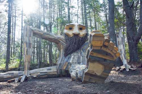 A large wooden sculpture of a giant figure with a beard, sitting in a forest surrounded by trees.