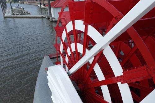 Close-up of a red and white paddle wheel on a riverboat, partially submerged in water with docks in the background.