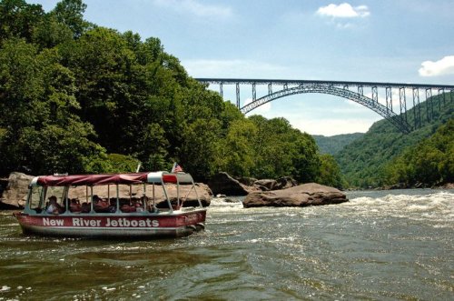 A jetboat navigates the New River, with a large bridge and lush green hills in the background under a clear blue sky.