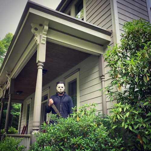 A figure in a mask and dark clothing stands on a porch, holding a weapon, surrounded by greenery.