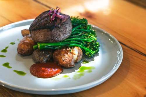 A gourmet plate featuring a grilled steak, broccolini, roasted potatoes, and a drizzle of sauce on a white dish.