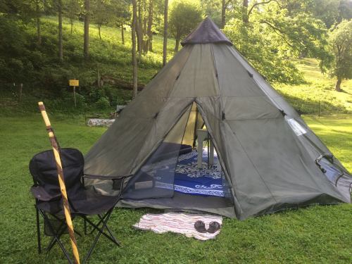 A tent set up on green grass, with a chair, walking stick, and a patterned mat outside in a wooded area.