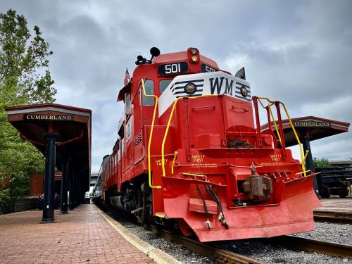 A red train engine at Cumberland station, with a cloudy sky in the background and historic architecture nearby.