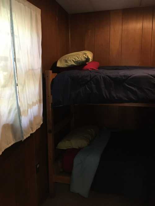 A cozy room with wooden walls featuring a bunk bed, colorful pillows, and a window with sheer curtains.