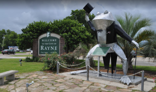 A large metal frog statue in front of a welcome sign for the city of Rayne, surrounded by greenery and a stone path.