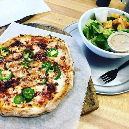 A pizza topped with jalapeños on a wooden board, alongside a salad with croutons and dressing on a plate.