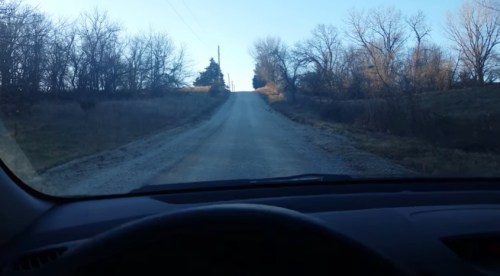 View from inside a car looking up a gravel road lined with trees, leading to a hill in the distance.