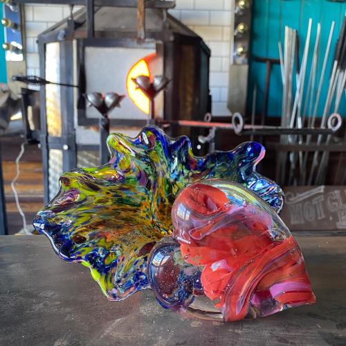 A colorful glass bowl and a heart-shaped glass piece on a workbench, with a glassblowing furnace in the background.