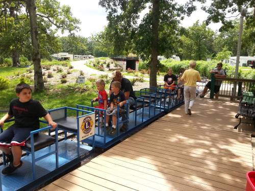 Children and adults sit on a small train ride in a park, surrounded by trees and greenery.