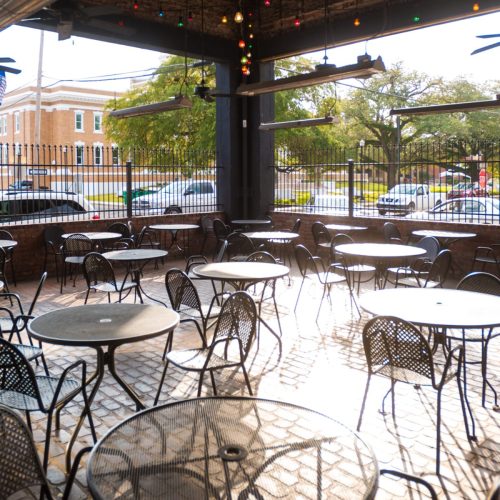Outdoor dining area with empty tables and chairs, surrounded by greenery and string lights.