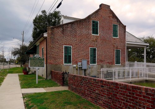 Historic brick house with green shutters, surrounded by a grassy area and a sidewalk, under a cloudy sky.