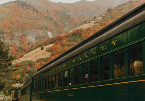 A vintage green train car in a scenic valley with colorful autumn foliage and mountains in the background.