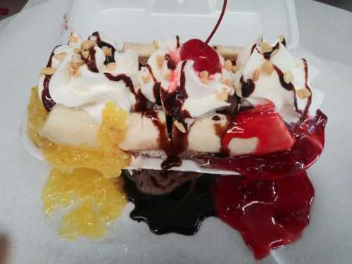 A banana split topped with whipped cream, cherries, nuts, and colorful jellies on a white takeout container.