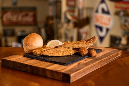A plate featuring two fried fish fillets, a lemon wedge, a roll, and two hushpuppies on a wooden board.