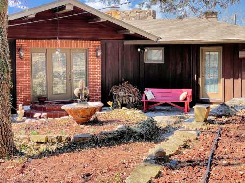 A cozy house with a wooden exterior, a red bench, and a stone fountain in the front yard, surrounded by greenery.
