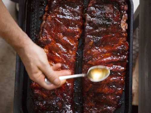 A hand drizzles sauce over two glazed, barbecued ribs on a black tray.