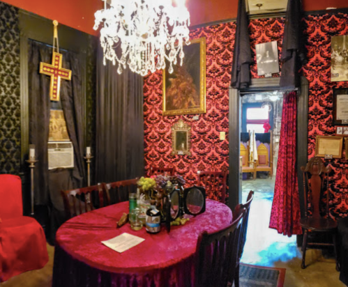 A dimly lit room with red and black decor, featuring a chandelier, a cross, and a round table set for dining.