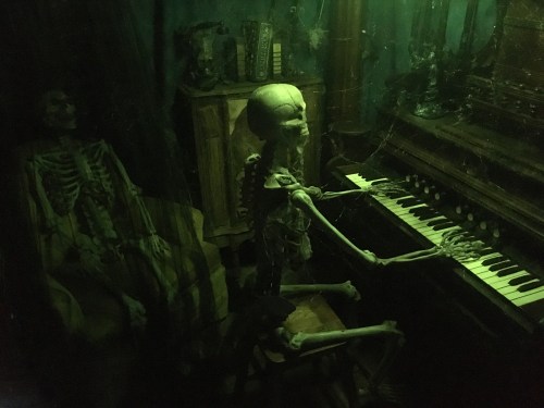 A dimly lit scene featuring two skeletons, one playing an organ and the other sitting in a chair nearby.
