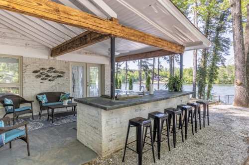 Outdoor bar area with seating, surrounded by trees and overlooking a lake. Bright and inviting atmosphere.