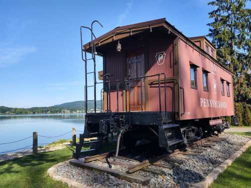 A vintage red caboose by a lake, surrounded by greenery and mountains in the background.