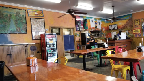 A colorful restaurant interior with wooden tables, bright chairs, and vibrant wall art. A staff member is seen in the background.
