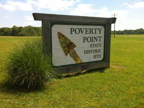 Sign for Poverty Point State Historic Site, featuring a spearhead design, set in a grassy field under a blue sky.