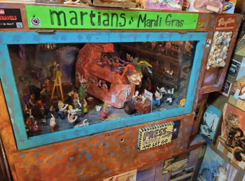 A colorful display featuring miniature Martians and Mardi Gras scenes inside a glass case with vibrant decorations.