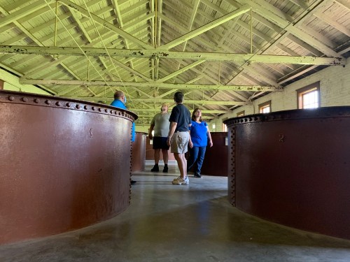 A group of four people stands in a large, industrial space with curved metal tanks and a high, wooden ceiling.