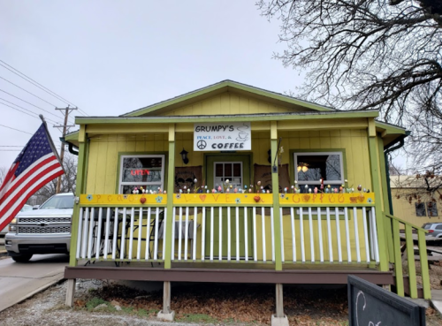 A yellow house with a porch, featuring a sign for "Grumpy's" coffee shop and an American flag in front.