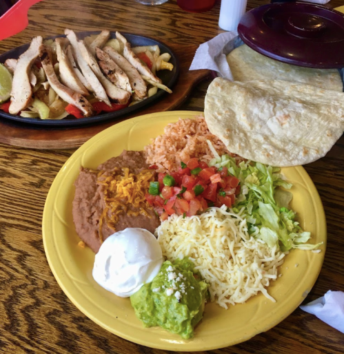 A colorful plate with refried beans, rice, lettuce, tomatoes, cheese, guacamole, sour cream, and tortillas, alongside grilled chicken.