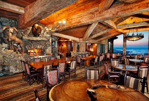 Cozy rustic dining room with wooden beams, stone fireplace, and large windows showcasing a scenic view.
