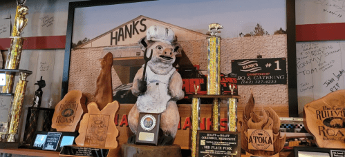 A wooden pig statue in front of a restaurant backdrop, surrounded by various trophies and awards.