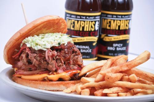 A hearty pulled pork sandwich topped with coleslaw, served with crispy fries and two bottles of barbecue sauce in the background.
