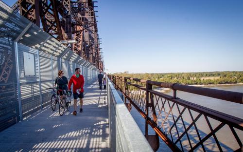 People walking and biking on a bridge with a metal structure, overlooking a river and green landscape.