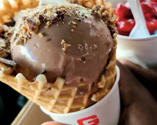 A close-up of a chocolate ice cream cone topped with crushed nuts, held in a hand, with a bowl of red toppings in the background.
