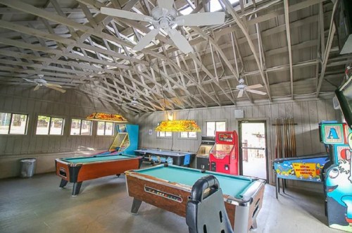 Interior of a game room featuring pool tables, arcade machines, and ceiling fans in a rustic setting.