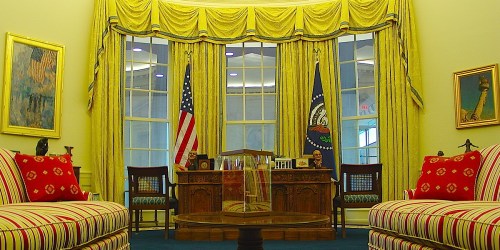 The Oval Office featuring yellow drapes, a wooden desk, and red-accented sofas, with flags and artwork in the background.