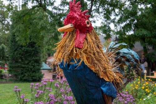 A colorful, large rooster sculpture made from various materials, surrounded by flowers and greenery.